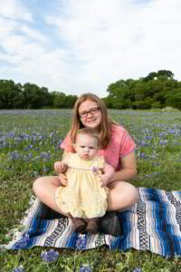A woman with a baby sitting on a blanket in a field of bluebonnets.